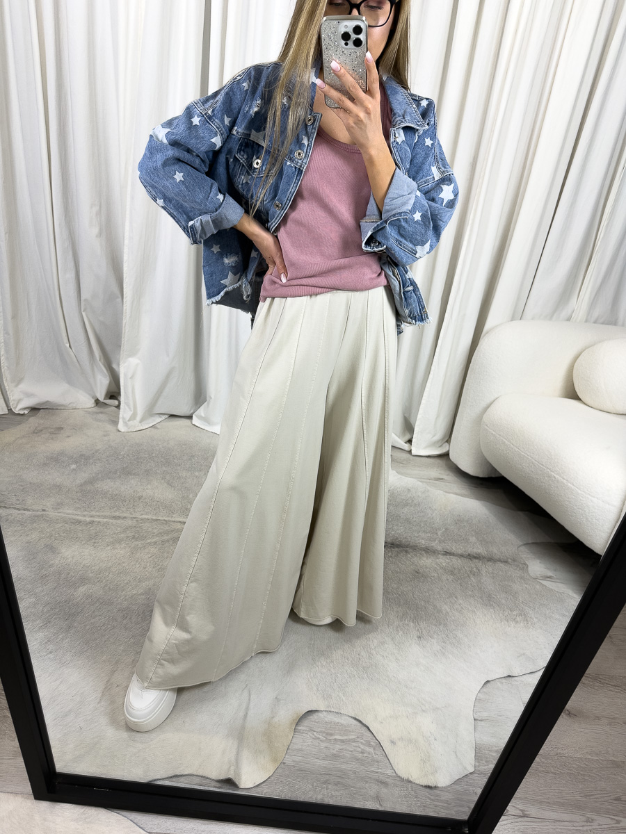 IMG_2083 Modelka prezentuje luźne spodnie Baggy Wiya w beżowym kolorze, z szerokimi nogawkami i wysokim stanem, zestawione z różowym topem i jeansową kurtką z gwiazdkami, w stylizacji casualowej na tle jasnych zasłon.