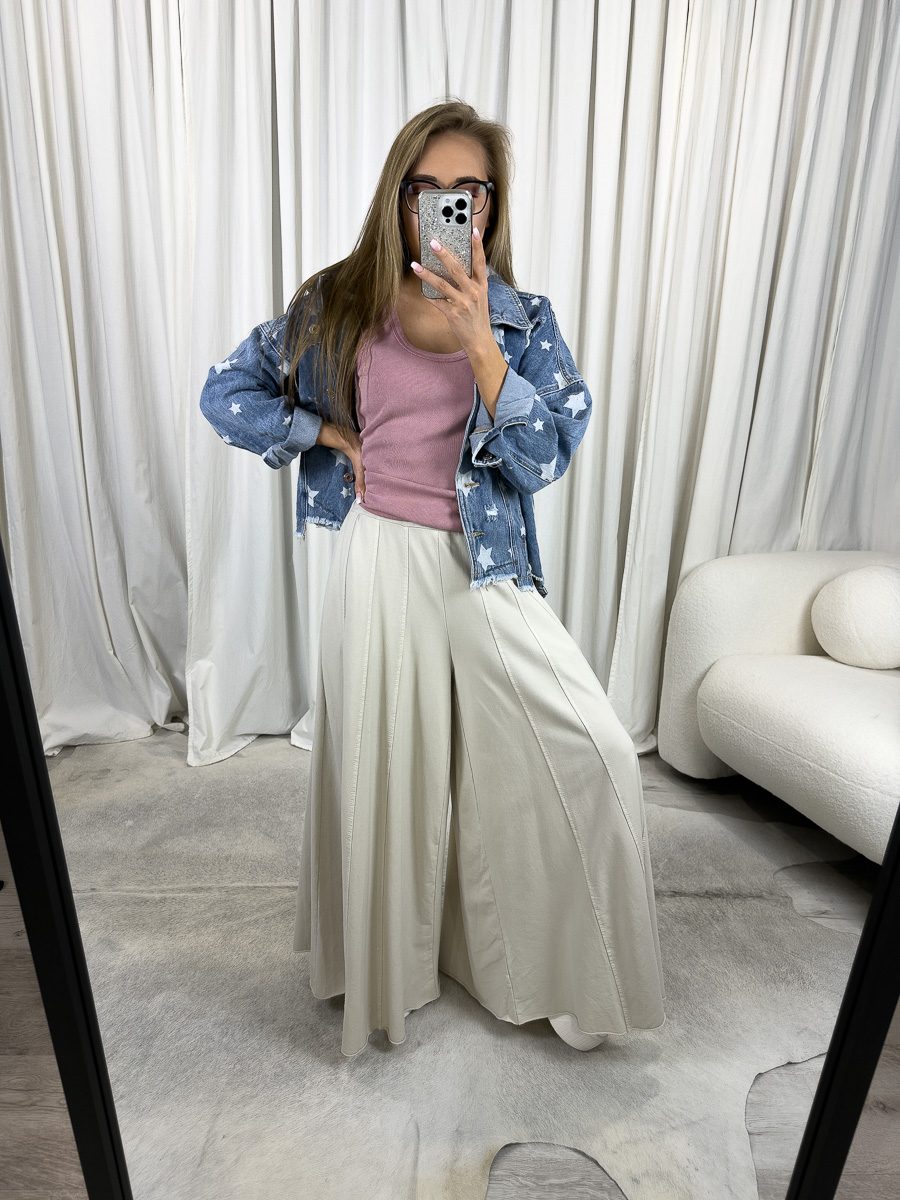 IMG_2093 Modelka prezentuje luźne spodnie Baggy Wiya w beżowym kolorze, z szerokimi nogawkami i wysokim stanem, zestawione z różowym topem i jeansową kurtką z gwiazdkami, w stylizacji casualowej na tle jasnych zasłon.