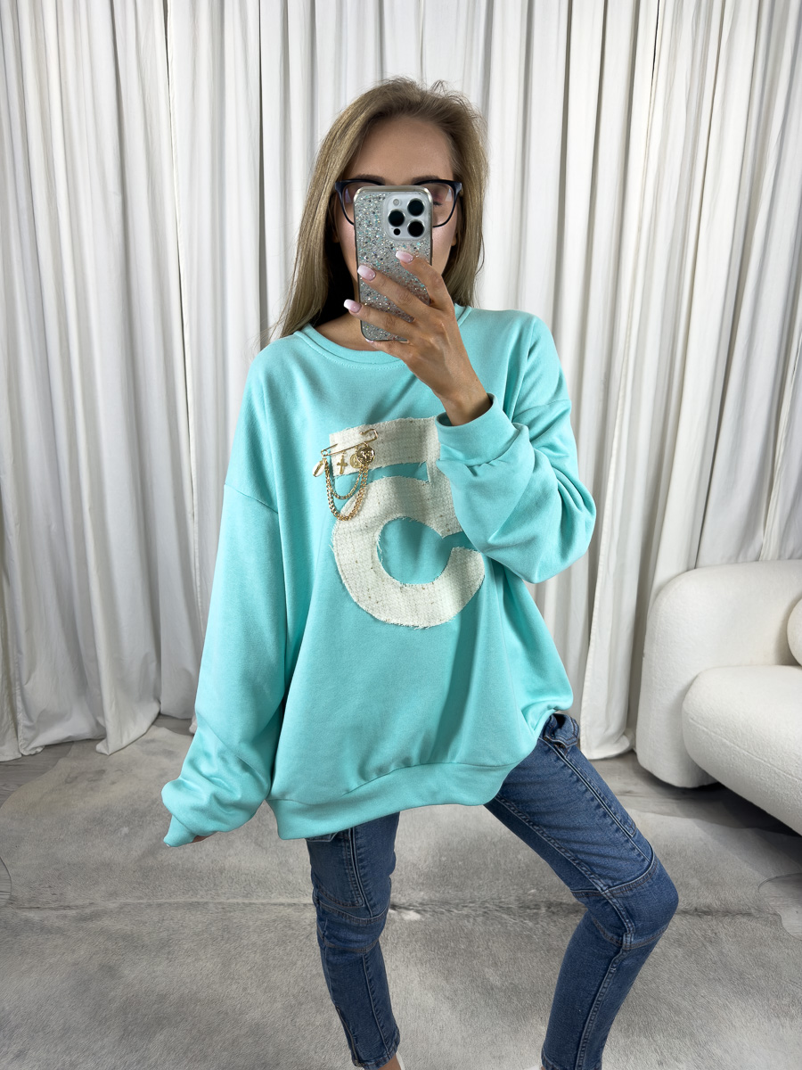 IMG_2600 Bluza oversize Five turkusowa z dużą cyfrą 5 i ozdobą, stylizacja z jeansami i białymi sneakersami, zdjęcie produktowe na tle jasnych zasłon.