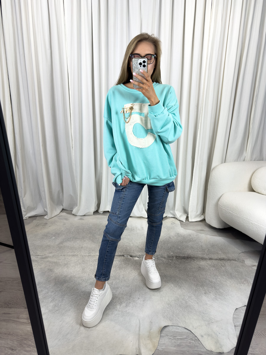 IMG_2607 Bluza oversize Five turkusowa z dużą cyfrą 5 i ozdobą, stylizacja z jeansami i białymi sneakersami, zdjęcie produktowe na tle jasnych zasłon.